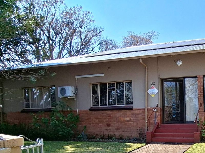 Front elevation of a single-story house featuring a brick and plaster facade, metal roof, security bars, and a paved walkway leading to the entrance.