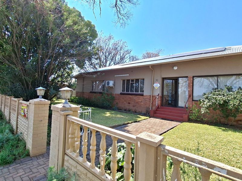 Front view of a single-story brick house featuring a paved walkway, front garden, boundary wall with balustrades, and roof-mounted solar panels.