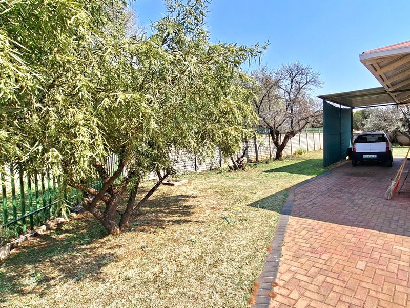 A spacious garden area featuring a paved driveway leading to a covered carport and enclosed by perimeter walling.