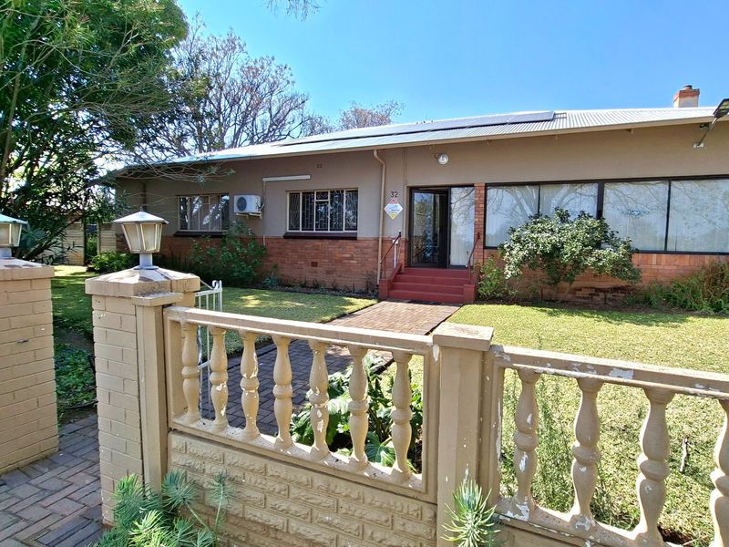 Front view of a single-story house featuring a brick boundary wall, paved entrance path, solar panels, and an established garden.