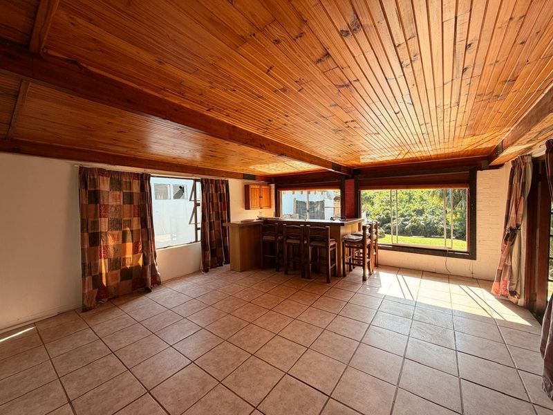 A spacious bar area featuring a built-in counter, warm timber tongue and groove ceiling, tiled flooring, and large windows overlooking the garden.