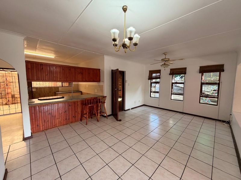 Spacious open-plan living and kitchen area featuring tiled flooring, wooden cabinetry, a breakfast bar, and ample natural light through multiple windows.