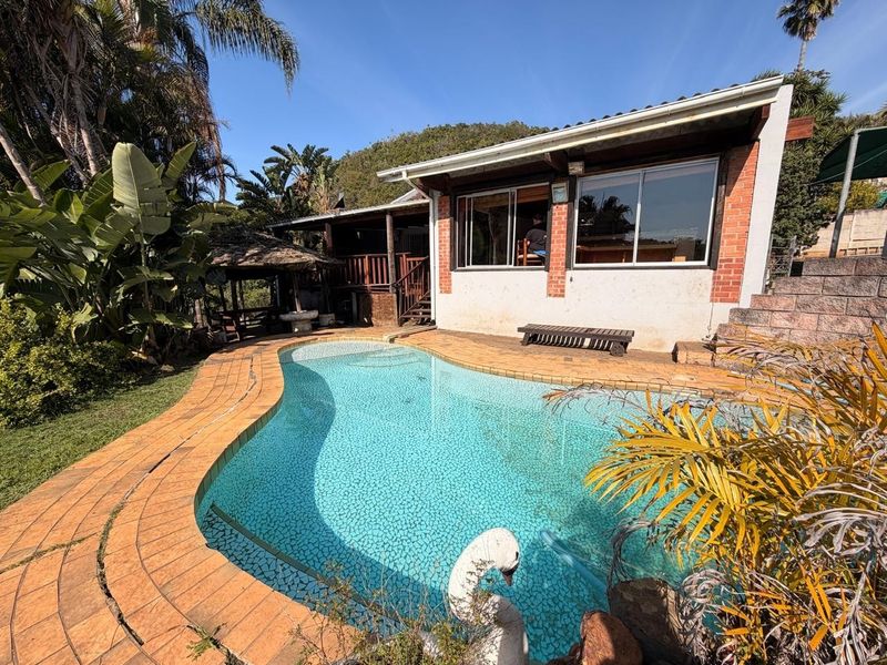 Outdoor pool area featuring a curved swimming pool with brick paving, a retaining wall, and a small thatched gazebo in the garden.