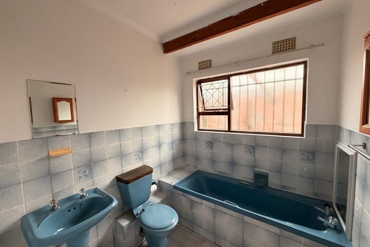 A dated bathroom featuring a blue pedestal basin, matching toilet, and built-in bathtub with tiled walls and an exposed timber ceiling beam.