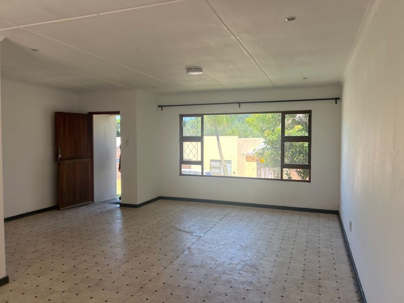 A spacious, empty living room featuring tiled flooring, a large window, and a timber entrance door, offering a blank canvas for renovation.
