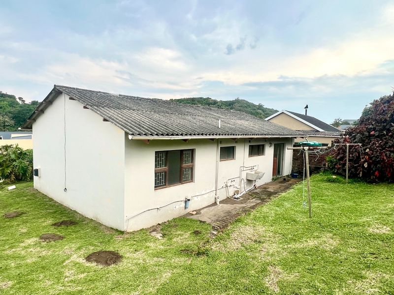 Rear view of the property showing the exterior wall, corrugated roof, security-barred windows, and an external utility sink area set within a grassed garden.