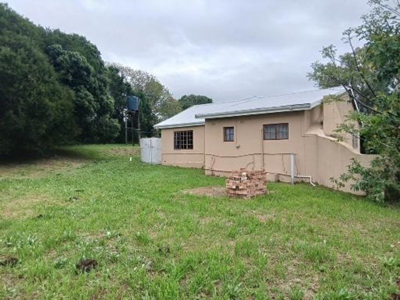 Rear view of a small holding dwelling featuring plastered masonry walls, a corrugated metal roof, and external water storage infrastructure set on a grassy plot.