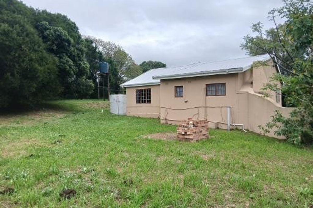 Rear view of a small holding dwelling featuring plastered masonry walls, a corrugated metal roof, and external water storage infrastructure set on a grassy plot.