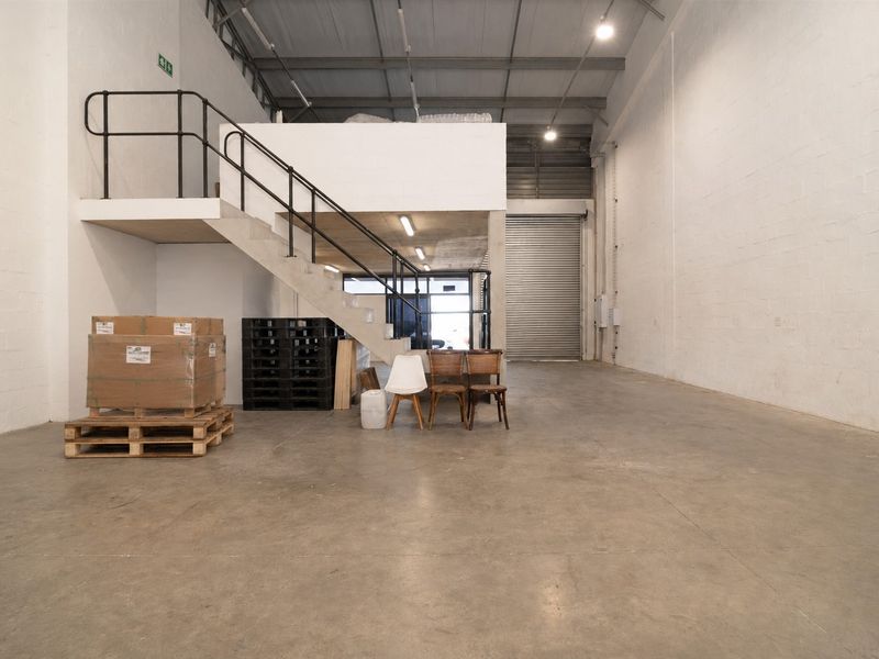 Spacious commercial warehouse unit featuring high ceilings, a mezzanine level with metal staircase, concrete flooring, and a large roller shutter door for easy access.
