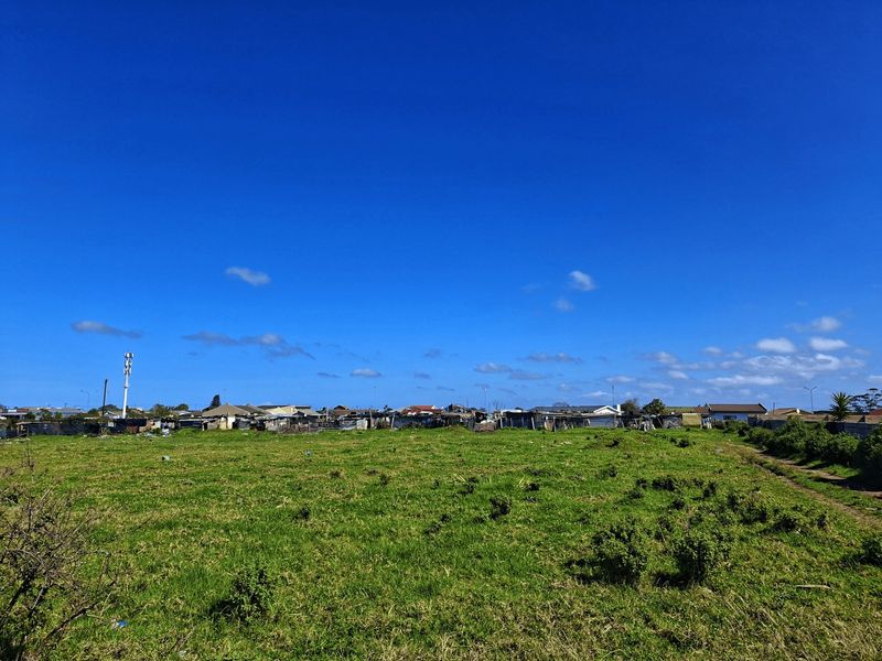 A wide-angle view of a vacant residential plot featuring open grassland with surrounding neighborhood structures in the distance.
