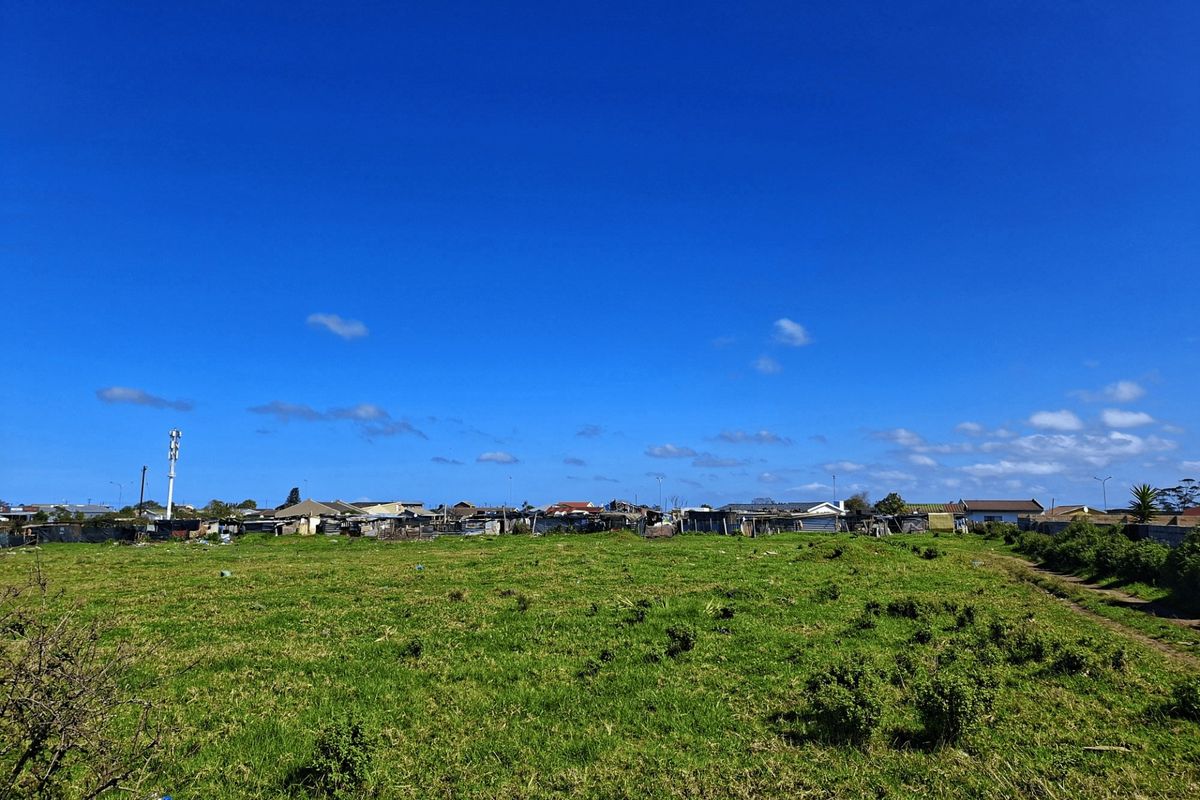 A wide-angle view of a vacant residential plot featuring open grassland with surrounding neighborhood structures in the distance.