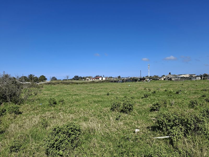 A wide-angle view of a vacant residential plot featuring open grassland with surrounding neighborhood infrastructure in the distance.