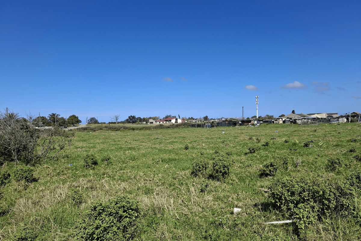 A wide-angle view of a vacant residential plot featuring open grassland with surrounding neighborhood infrastructure in the distance.