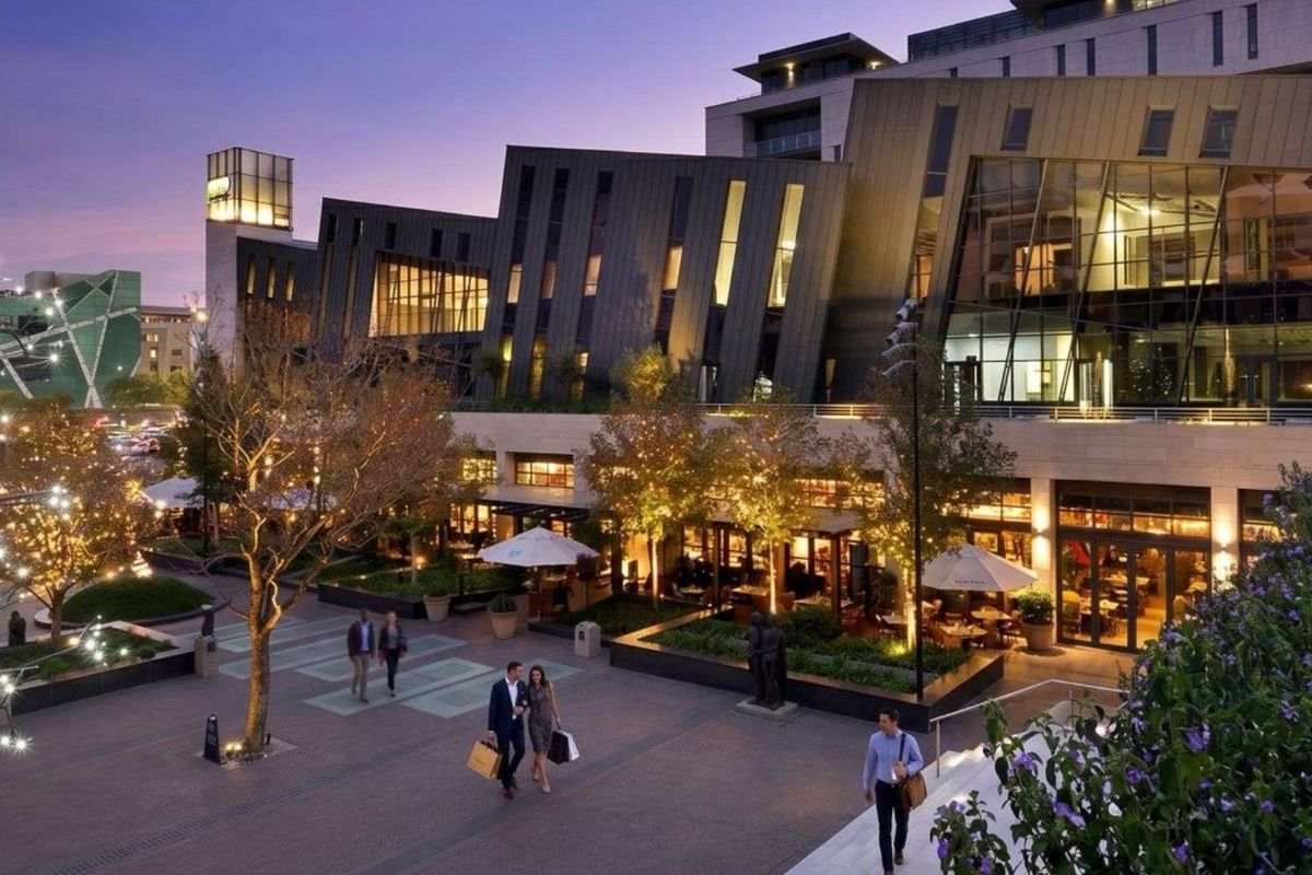 A contemporary apartment building exterior featuring a modern glass and metal facade overlooking a beautifully landscaped, paved courtyard area at dusk.