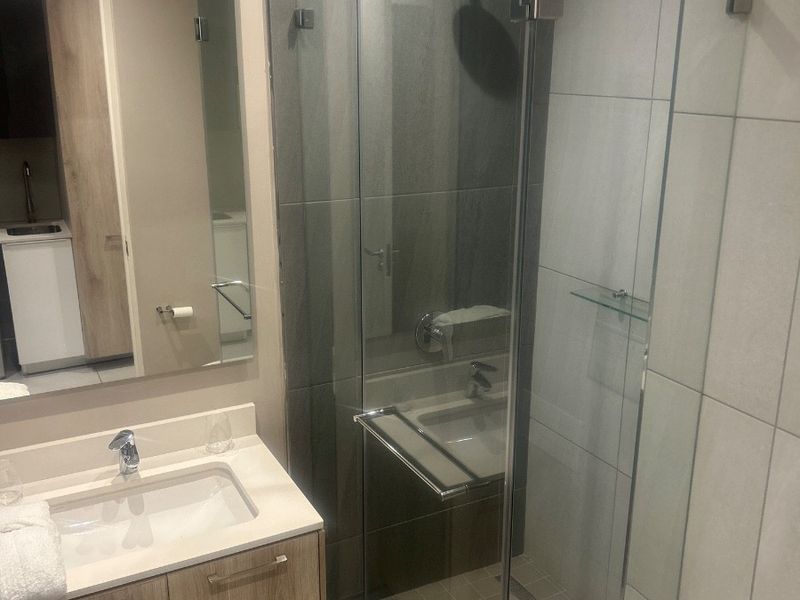 Modern bathroom featuring a wood-finish vanity with integrated sink, wall-mounted mirror, and a spacious glass-enclosed shower with contemporary tiling.