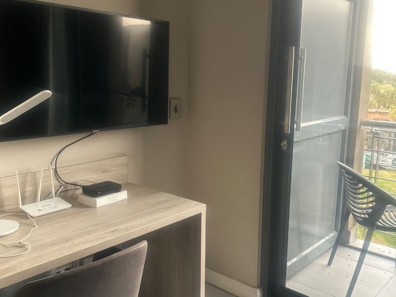 A functional office space featuring tiled flooring, a wall-mounted television, and direct access to a private balcony through a sliding glass door.