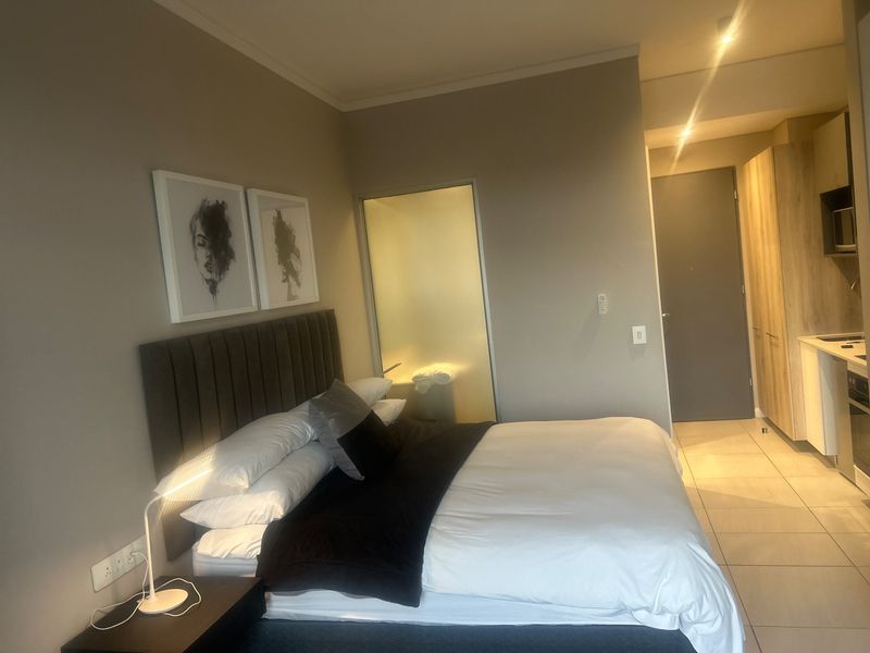 A modern bedroom featuring tiled flooring, recessed lighting, and a frosted glass door leading to an en-suite bathroom, with adjacent built-in kitchen cabinetry.