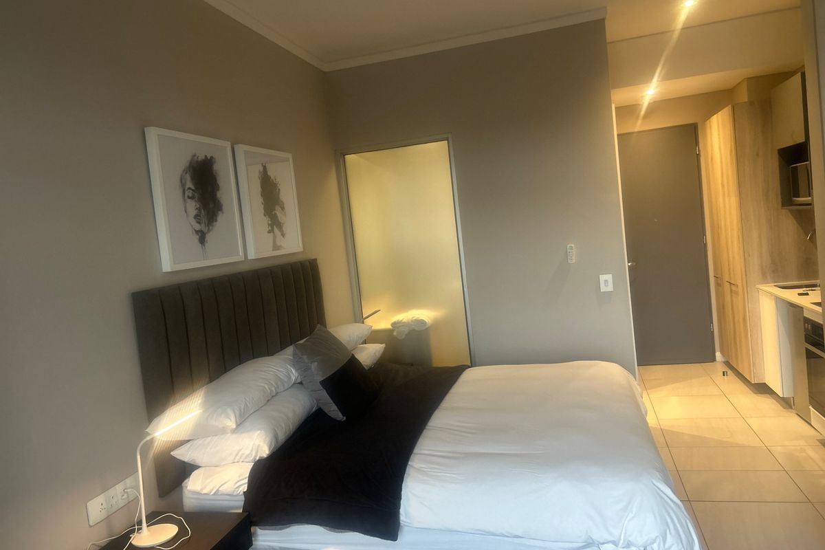 A modern bedroom featuring tiled flooring, recessed lighting, and a frosted glass door leading to an en-suite bathroom, with adjacent built-in kitchen cabinetry.