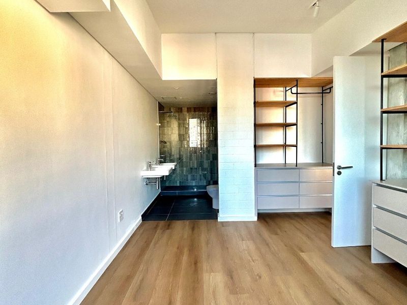 Modern bedroom featuring light wood-look flooring, built-in open shelving, integrated cabinetry, and an open-plan en-suite bathroom with a tiled shower and wall-mounted vanity.