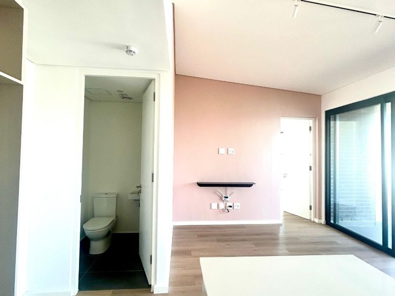 A bright, modern bedroom featuring laminate flooring, track lighting, a private en-suite bathroom, and large sliding glass doors leading to an outdoor area.