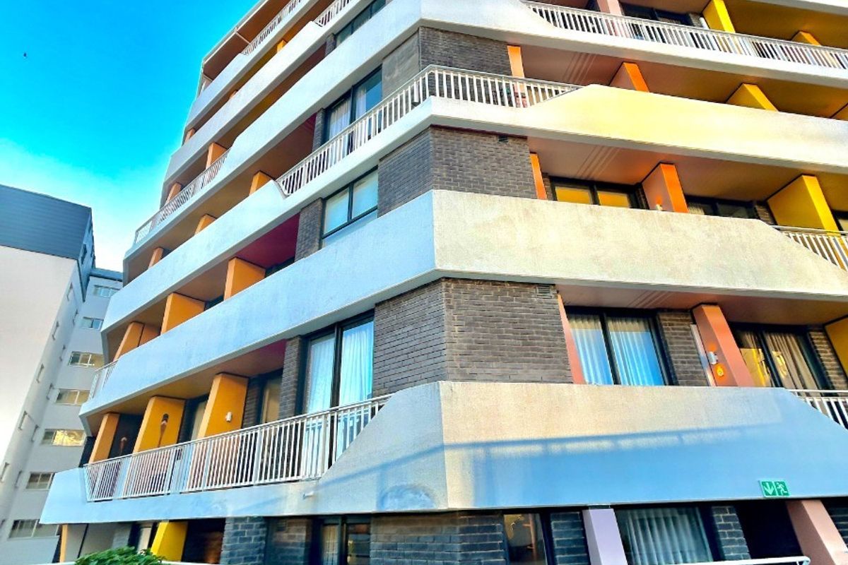 A low-angle view of a multi-story apartment building featuring brick facade, private balconies with metal railings, and large sliding glass doors.