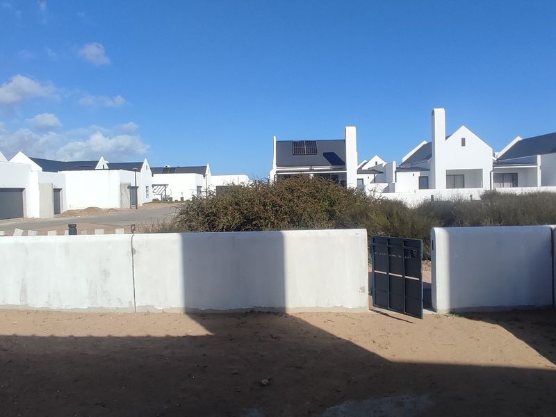 A view of the rear exterior area featuring a boundary wall, pedestrian gate, and unlandscaped sandy ground in a residential estate setting.