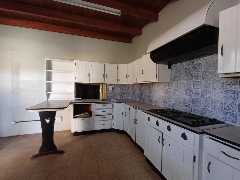 A dated kitchen featuring white cabinetry, patterned tile backsplash, integrated hob with extractor, and exposed timber ceiling beams.