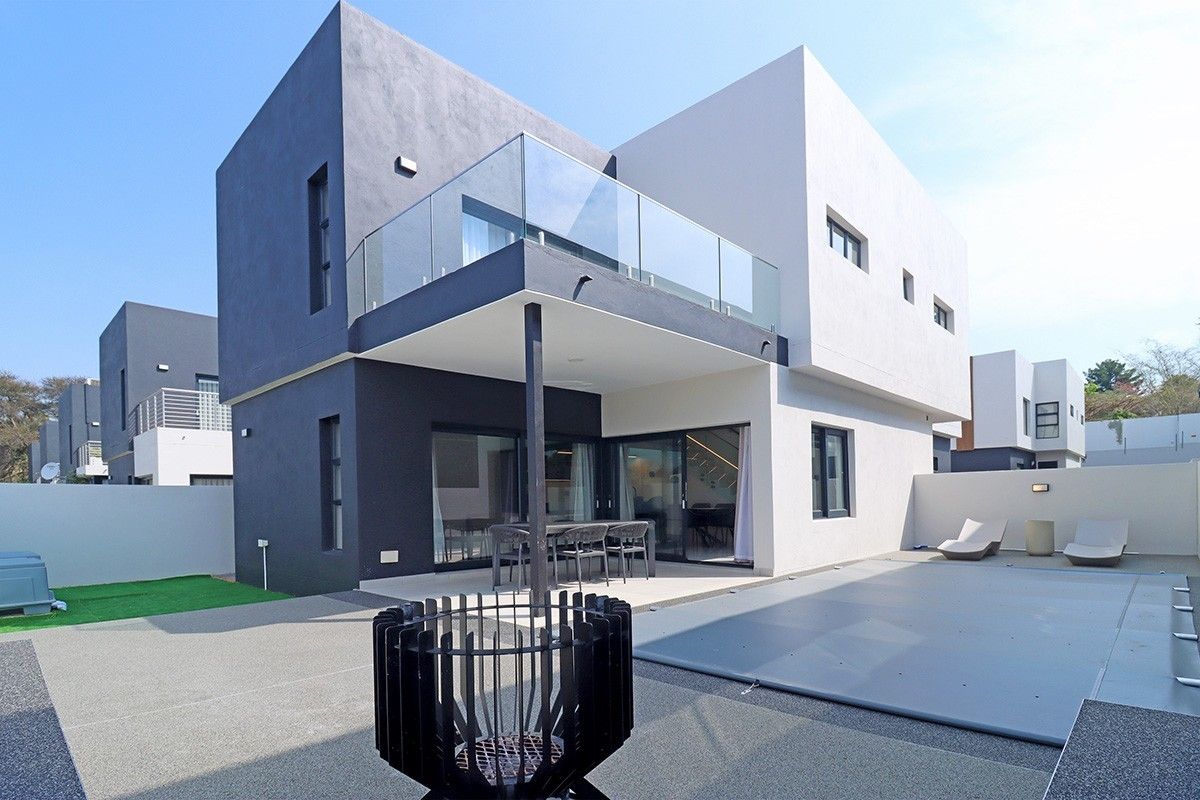 Modern double-story rear elevation featuring a covered patio, glass balcony, paved terrace, and a swimming pool with a safety cover.