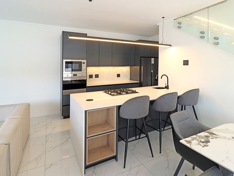 Modern open-plan kitchen featuring a central island with gas cooktop, sleek dark cabinetry, integrated appliances, and elegant marble-look floor tiles.
