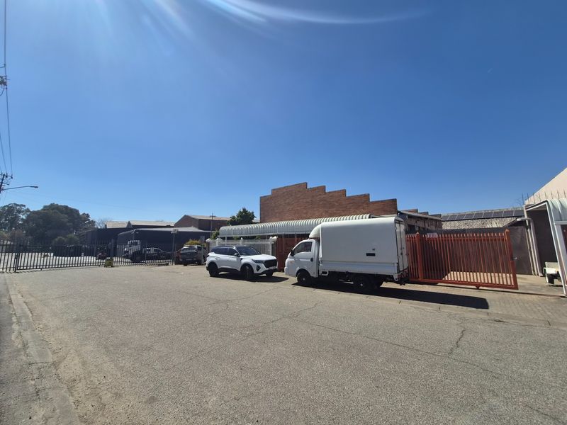Front exterior view of an industrial property featuring a secure gated entrance, asphalt driveway, and brick facade with corrugated metal roofing.