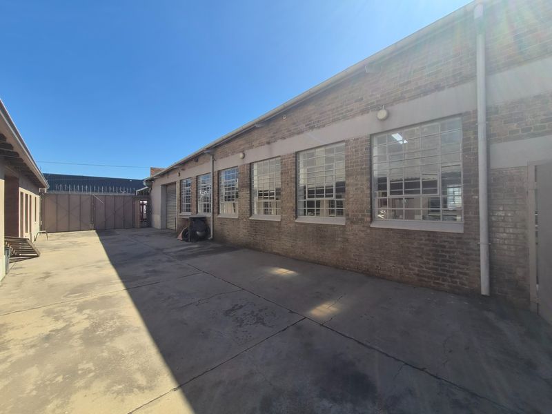 Rear industrial yard featuring exposed brick walls, large steel-framed windows with security bars, and a concrete surface with a roller shutter door access.