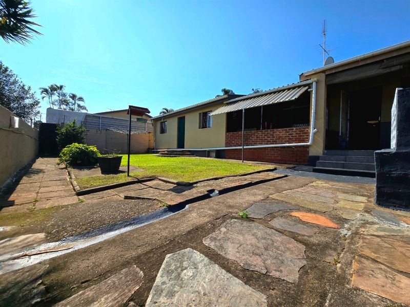 Rear exterior view of the townhouse featuring a paved patio area, a small lawn, and a covered outdoor space with a brick retaining wall.