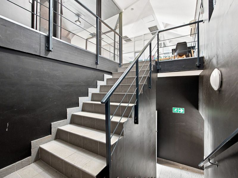 A modern office hallway featuring a tiled staircase with metal balustrades and glass partitions, leading to an upper mezzanine level.