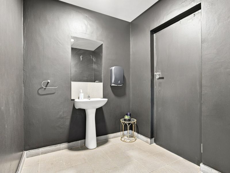 A functional office restroom featuring a pedestal sink, wall-mounted mirror, and paper towel dispenser with dark painted walls and tiled flooring.