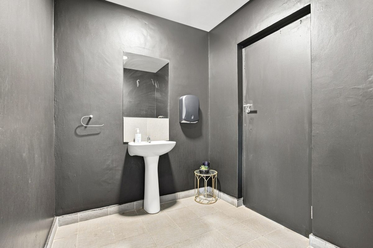 A functional office restroom featuring a pedestal sink, wall-mounted mirror, and paper towel dispenser with dark painted walls and tiled flooring.