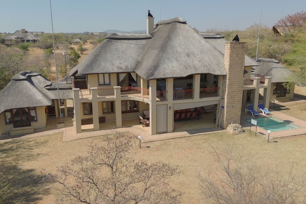 Aerial view of a large thatched-roof house featuring a swimming pool, expansive covered patio, and balcony, set within a natural bushveld landscape.