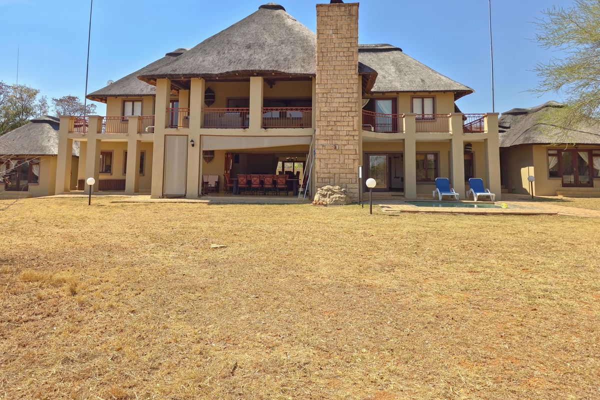 Rear view of a large double-story house featuring a traditional thatch roof, stone chimney, expansive covered patio, and a private swimming pool.