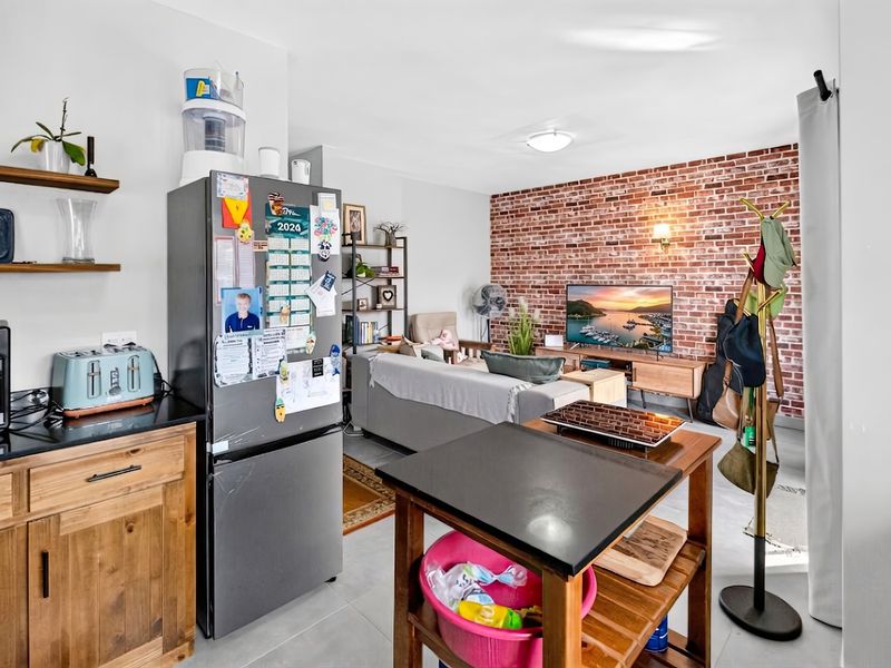 Open plan living area featuring an exposed brick feature wall, tiled flooring, and a compact kitchen area with wooden cabinetry and stone countertops.