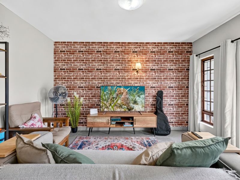 A well-lit living room featuring a prominent exposed brick feature wall, tiled flooring, and a window with security bars.