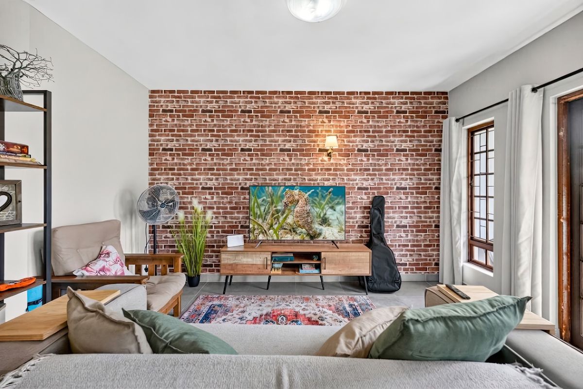 A well-lit living room featuring a prominent exposed brick feature wall, tiled flooring, and a window with security bars.