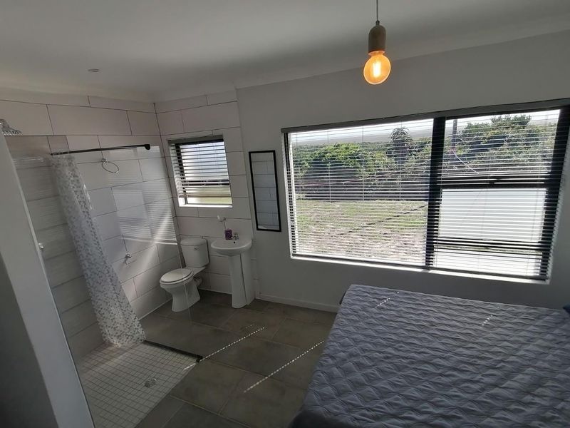 A modern bedroom featuring an open-plan en-suite bathroom with a walk-in shower, pedestal basin, and large windows providing natural light.