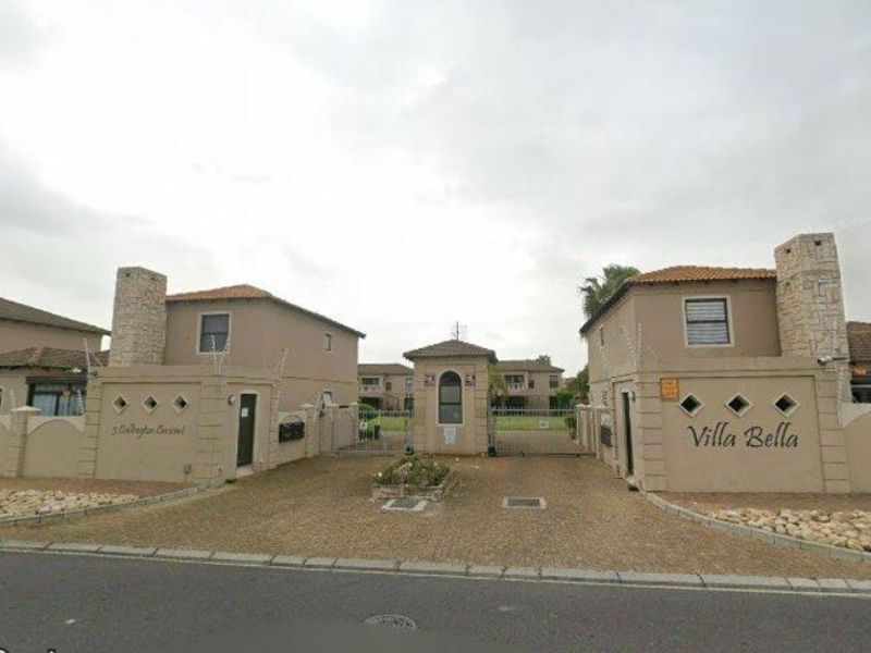 The gated entrance to Villa Bella townhouse complex, featuring stone-clad pillars, security fencing, and a paved driveway approach.