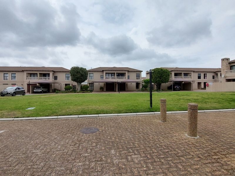 A view of the townhouse complex exterior featuring a paved driveway, communal lawn area, and multiple residential units with balconies and carports.