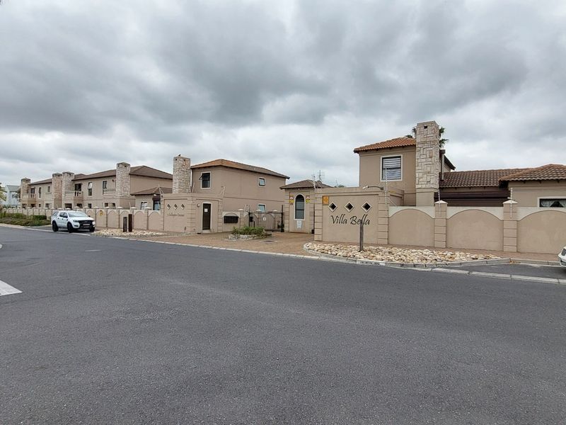 Street view of the Villa Bella townhouse complex entrance, featuring perimeter walls with stone accents and tiled roofing under an overcast sky.