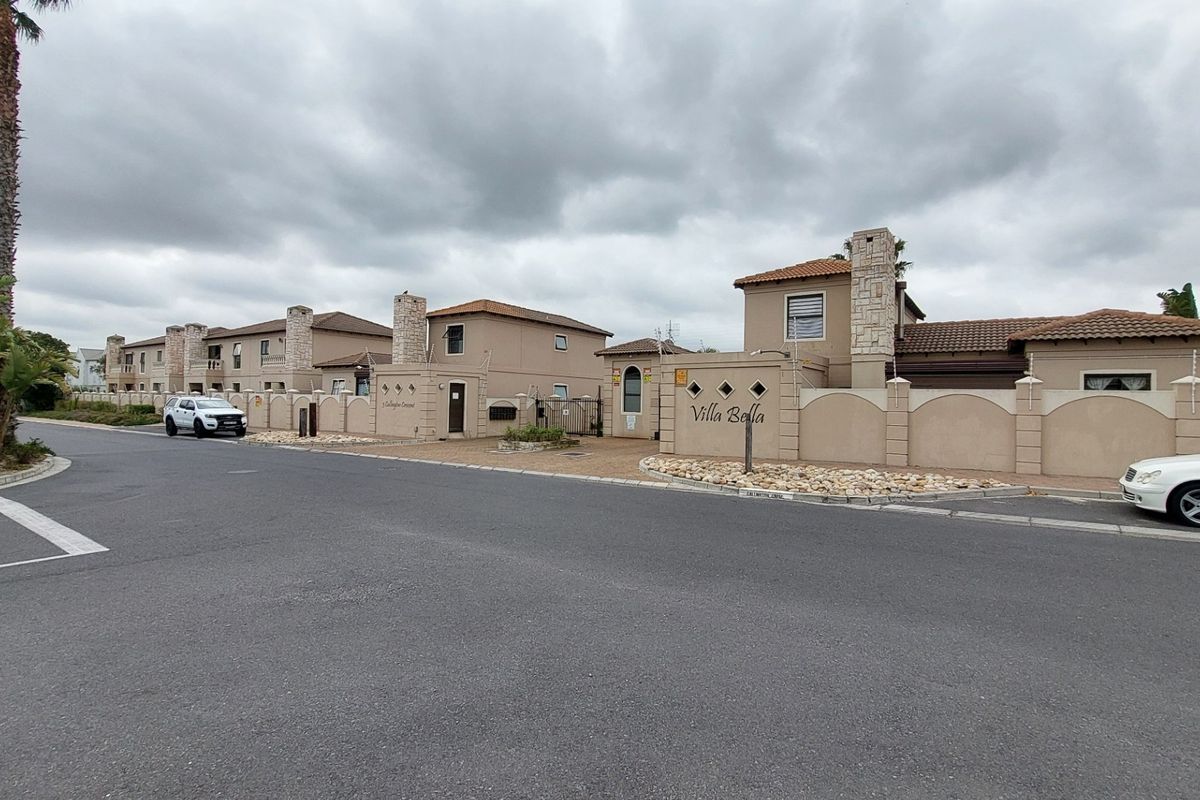 Street view of the Villa Bella townhouse complex entrance, featuring perimeter walls with stone accents and tiled roofing under an overcast sky.