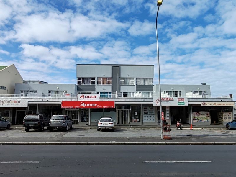 Front exterior view of a multi-level commercial apartment building featuring ground-floor retail storefronts, upper-level balconies, and a paved street-level parking area.