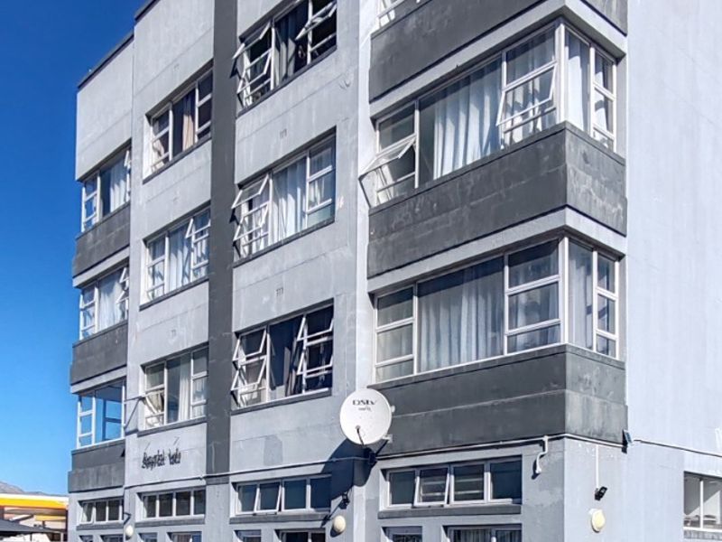 A multi-story apartment building exterior featuring a grey facade, multiple casement windows, and dedicated ground-level parking bays.