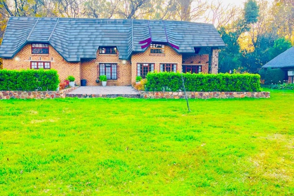 Rear view of a facebrick small holding residence featuring a thatched roof, stone retaining wall, and a paved patio area overlooking a large lawn.