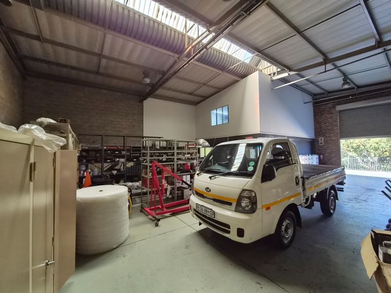 Spacious industrial warehouse or garage featuring high ceilings with skylights, exposed steel trusses, concrete flooring, and a large roller shutter door for vehicle access.