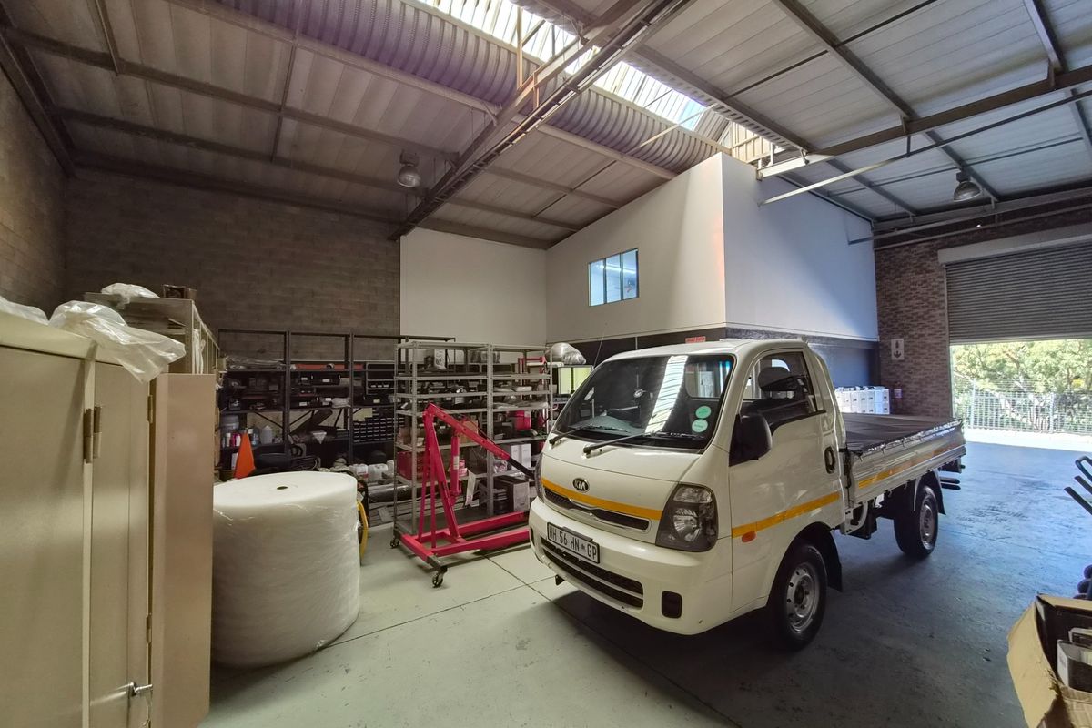 Spacious industrial warehouse or garage featuring high ceilings with skylights, exposed steel trusses, concrete flooring, and a large roller shutter door for vehicle access.